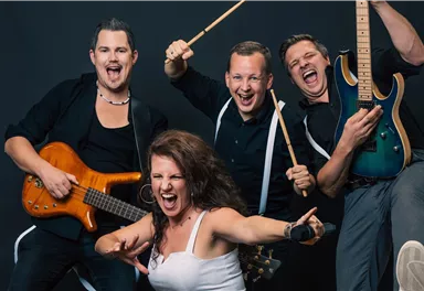 Eine lebendige Band mit vier Mitgliedern posiert enthusiastisch für das Foto. Sie halten Musikinstrumente und zeigen Freude und Energie auf der Bühne.