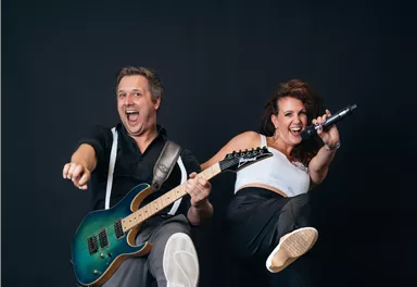 Ein Musiker mit einer Gitarre und eine Sängerin posieren fröhlich vor einem neutralen Hintergrund. Beide strahlen Energie und Freude aus.