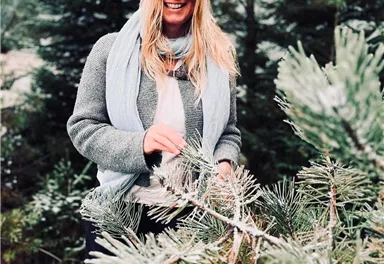 A smiling woman stands among green fir trees. She is wearing a gray sweater and a scarf and is enjoying nature.
