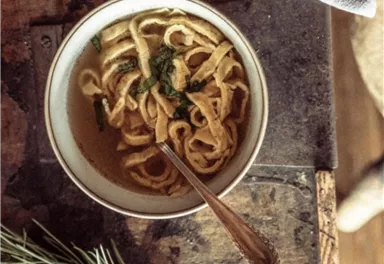 A bowl of homemade noodle soup is sitting on a wooden table. Next to it are a few pine branches and a gray cloth.