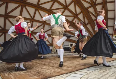 Eine Gruppe von Tänzern in traditioneller bayerischer Tracht tanzt auf einer Bühne. Die Frauen tragen lange schwarze Röcke und die Männer kurze Hosen mit Socken.