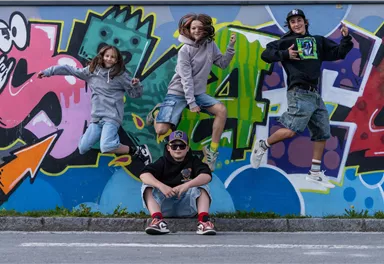 Vier Kinder springen vor einer bunten Graffiti-Wand. Sie haben Spaß und tragen sportliche Kleidung.