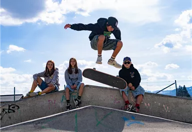 Eine Gruppe von Jugendlichen beim Skateboarden im Skatepark. Ein Junge macht einen Sprung über das Skateboard, während die anderen zuschauen.