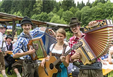 Eine fröhliche Gruppe von Musikern spielt auf einem Fest im Freien. Sie tragen traditionelle Trachten und sind umgeben von einer malerischen Waldlandschaft.