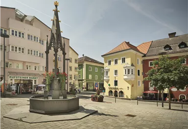 Ein schöner Platz mit einem Brunnen in der Mitte und bunten Gebäuden im Hintergrund. Die Atmosphäre ist ruhig und einladend.