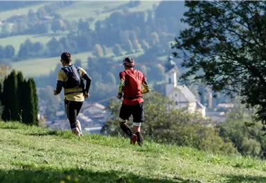 Zwei Läufer joggen über eine grüne Wiese. Im Hintergrund sind sanfte Hügel und ein kleiner Ort mit einer Kirche zu sehen.