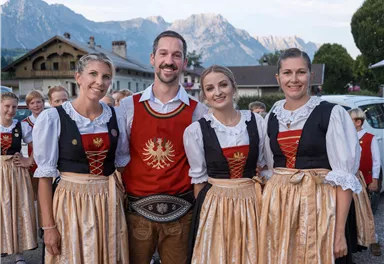 Eine Gruppe von vier Personen in traditioneller Tracht lächelt in die Kamera. Im Hintergrund sind Berge und eine ländliche Umgebung zu sehen.