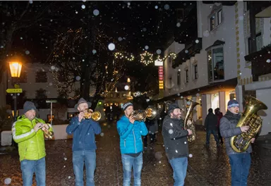 Eine Gruppe von Musikern spielt im Schneefall auf einer festlich beleuchteten Straße. Die Atmosphäre ist gemütlich und weihnachtlich.