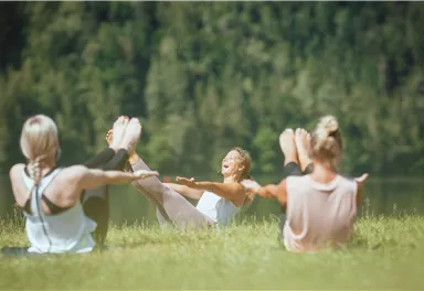 A group of women is doing yoga outdoors. They are sitting on a meadow with a view of a forest and a calm lake.