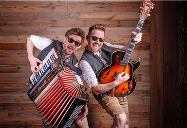 Zwei Männer, die fröhlich Musik spielen, stehen vor einer Holzwand. Einer spielt das Akkordeon, während der andere eine Gitarre hält und beide tragen coole Sonnenbrillen.