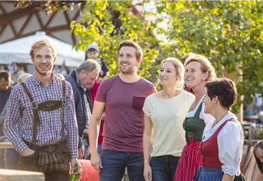 Eine Gruppe von fünf Menschen lächelt und genießt die Zeit im Freien. Sie tragen traditionelle Kleidung und stehen in einer festlichen Umgebung.
