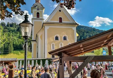 Ein lebhafter Marktplatz mit vielen Menschen und einer schönen, gelben Kirche im Hintergrund. Die Umgebung ist von grünen Hügeln und blauem Himmel umgeben.