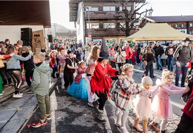 Eine fröhliche Kinderparade mit vielen bunt verkleideten Kindern, die eine Reihe bilden und tanzen. Im Hintergrund sind Zuschauer und Stände zu sehen, die zu einem Fest gehören.