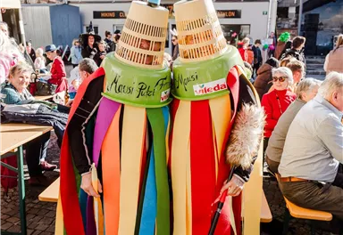 Zwei Personen in bunten Kostümen und Masken stehen nebeneinander. Im Hintergrund sind viele Menschen und eine festliche Atmosphäre zu sehen.
