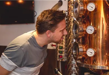 A man stands in front of a shiny copper still and is attentively looking at the measuring instruments. In the background, a television and industrial equipment can be seen.