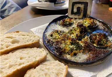 Ein Teller mit gebackenen Schnecken und knuspriger Kruste, serviert auf einem schwarzen Teller. Dazu liegen frische Brotscheiben.