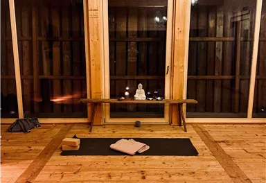 A quiet room with a wooden floor and large windows. In the center lies a yoga mat, surrounded by relaxing decoration.