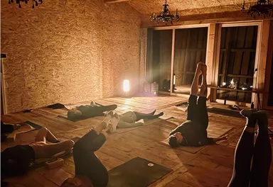 A relaxing yoga session in a cozy room with wooden ceilings and soft lighting. The participants are lying on mats and practicing various positions.