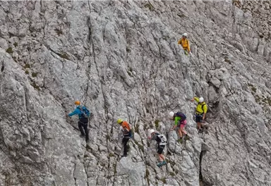 Eine Gruppe von Kletterern bewegt sich entlang einer Felswand. Sie tragen Helme und Kletterausrüstung, während sie eine steile Strecke überwinden.