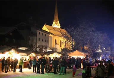 Ein festlicher Weihnachtsmarkt bei Nacht, beleuchtet mit bunten Lichtern. Menschen versammeln sich um Buden und genießen die Atmosphäre.