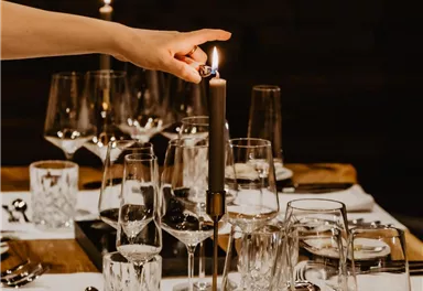 Ein elegant gedeckter Tisch mit Gläsern und Besteck. Eine Hand zündet eine Kerze an, die für eine stimmungsvolle Atmosphäre sorgt.