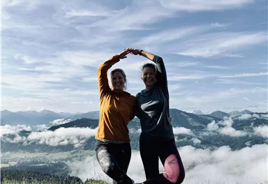 Zwei Frauen stehen auf einer grünen Wiese und formen mit ihren Armen ein Herz. Im Hintergrund sind Berge und Wolken zu sehen.