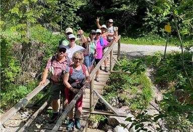 Eine Gruppe von Menschen überquert eine Holzbrücke in einer grünen Landschaft. Sie haben Hüte auf und genießen die Natur.