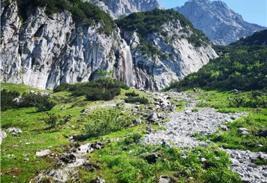 Ein malerischer Wasserfall, der von steilen Felsen umgeben ist. Die grüne Landschaft und die Berge im Hintergrund schaffen eine ruhige Atmosphäre.