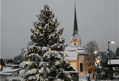 A festively decorated Christmas tree in front of a snow-covered church. The sky is gray and the surroundings feel wintry and cozy.