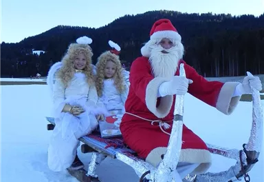 A Christmas sleigh with Santa Claus and two angels on a snowy field. In the background, there are forested hills and a clear blue sky.
