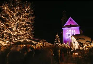 Ein festlicher Weihnachtsmarkt bei Nacht mit hell erleuchteten Ständen. Im Hintergrund ist eine Kirche mit lila Beleuchtung und ein geschmückter Weihnachtsbaum zu sehen.