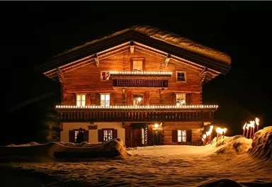 A beautiful wooden house in the snow, festively decorated with lights. Torches illuminate the path to the entrance, creating a warm atmosphere at night.
