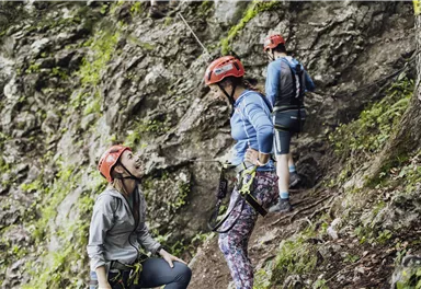 Three people in climbing gear stand at a rock wall. They are wearing helmets and seem to be discussing a climbing route.