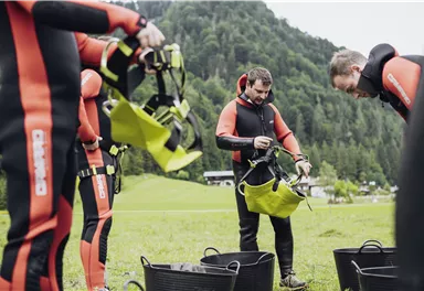 A group of people in wetsuits is preparing for an outdoor activity. Some participants are organizing their gear on a meadow amidst nature.