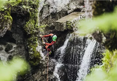A climber in a green forest is climbing a rock next to a waterfall. He is wearing a green helmet and using a rope for safety.