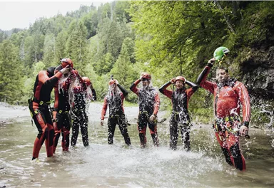 A group of seven people in wetsuits stands in the water and splashes with water. They are having a lot of fun in a natural setting with trees in the background.