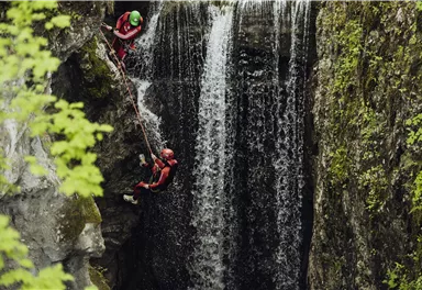 Two people are climbing a rock wall next to a waterfall. The surroundings are green and natural.