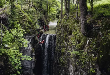 An impressive waterfall, surrounded by green trees, flows through a narrow gorge. Some people are active near the water.