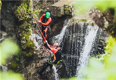 Two people in wetsuits climbing a rock wall. In the background, a waterfall is visible.