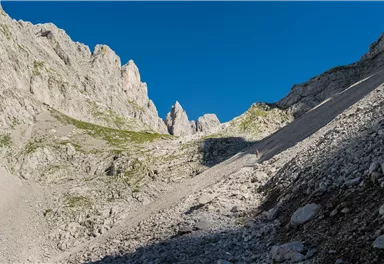 Eine beeindruckende Berglandschaft mit steilen Felswänden und einem klaren blauen Himmel. Der Weg ist von Geröll und Gras gesäumt.