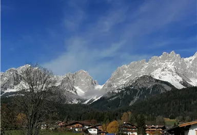 An impressive mountain landscape with snow-capped peaks and a clear blue sky. In the foreground, some houses and trees can be seen.