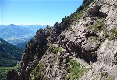A group of hikers is walking along a narrow path beside steep cliffs. In the background, a breathtaking mountain landscape stretches out.