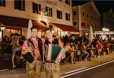 Two musicians are smiling on the street, one with a trumpet and the other with an accordion. In the background, many people can be seen in a lit restaurant and on the terrace.