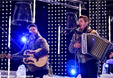 Two musicians perform on a illuminated stage. One plays guitar and the other an accordion.