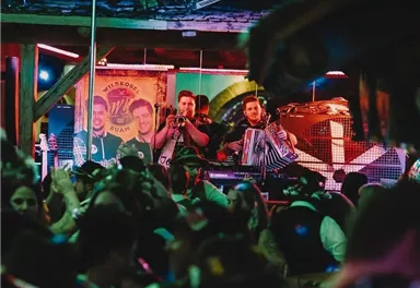 A vibrant party scene with many people and musicians on stage. Colorful lights and a carefree atmosphere characterize the surroundings.