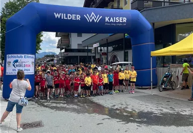 A group of children stands under a large blue start arch that bears the inscription "Wilder Kaiser." In the background, buildings and a yellow tent station are visible.