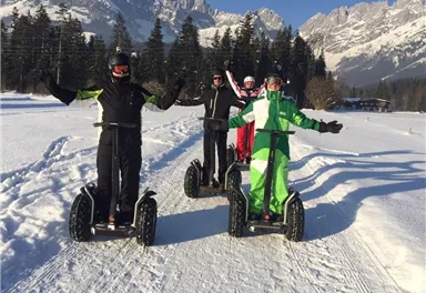 Four people are riding Segways through a snowy landscape. In the background, mountains and forests are visible.