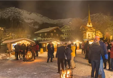 A wintry Christmas market at night with festively illuminated stalls. People enjoy the atmosphere in a snow-covered setting, surrounded by mountains.