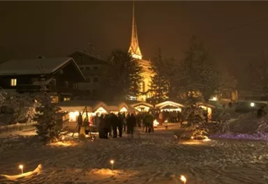 A winter market square with festive stalls and people at night. The snow and the illuminated church create a cozy atmosphere.