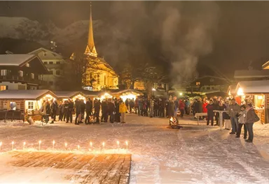 A winter market at night with many people. The festively lit stalls and the church in the background create a cozy atmosphere.